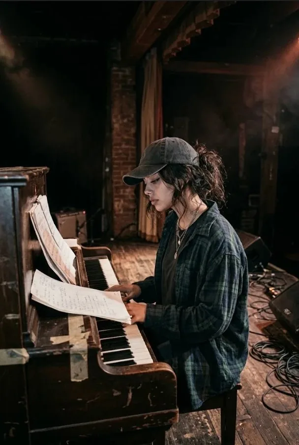 Vee Valentine at an upright piano in a ball cap, working through handwritten sheet music